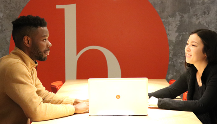 Two people sitting at a table with the bswift logo on the wall behind them