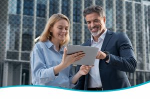 Two adults stand outdoors in front of a modern glass building with a circular pattern, jointly holding and viewing a tablet device; both wear business-casual clothing, and a curved teal graphic border appears along the bottom edge of the image.