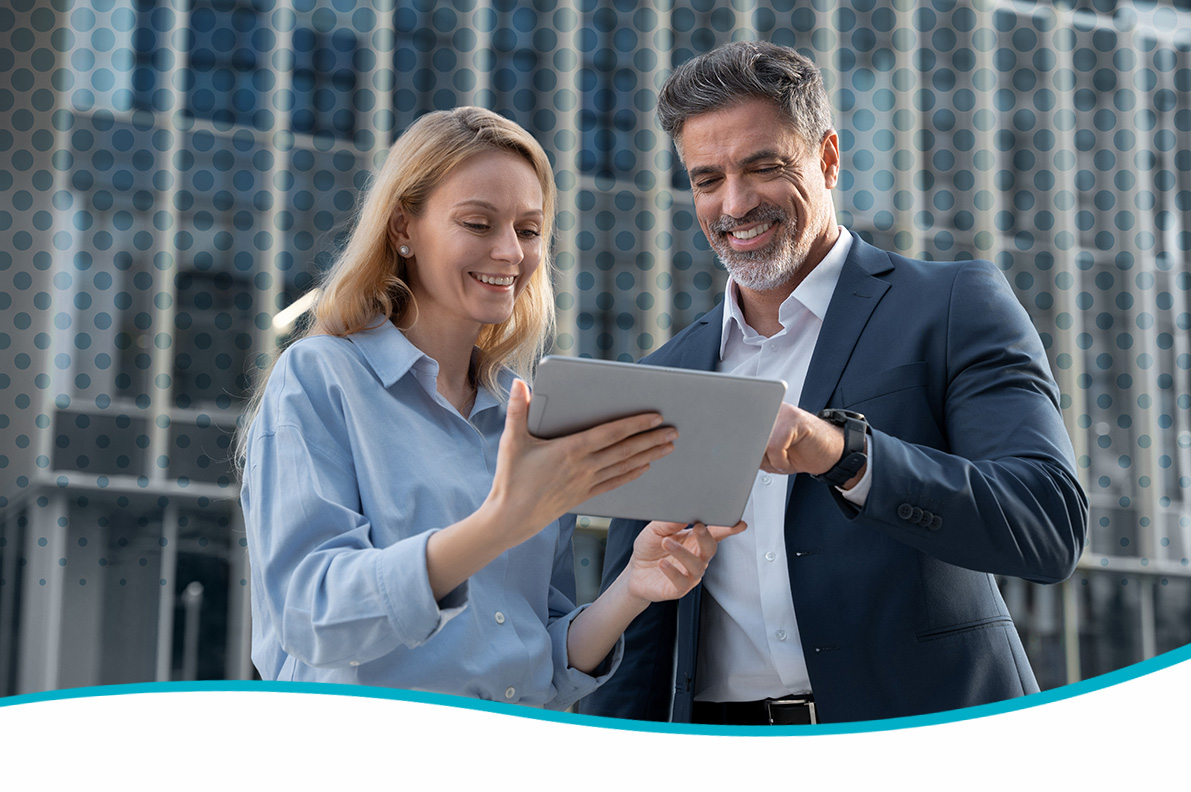 Two adults stand outdoors in front of a modern glass building with a circular pattern, jointly holding and viewing a tablet device; both wear business-casual clothing, and a curved teal graphic border appears along the bottom edge of the image.
